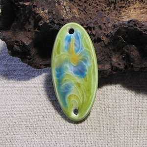 Può includere: Ciondolo ovale in ceramica con un motivo a iris dipinto. Il ciondolo presenta uno sfondo verde chiaro con un fiore di iris blu e giallo. Ha due fori per l'infilatura ed è lungo circa 5 cm.