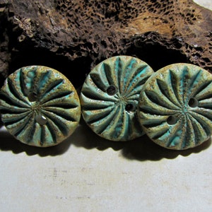 Op de afbeelding: Drie ronde keramische knopen in turquoise en bruin. Elke knoop heeft een radiaal ontwerp en twee gaten om te naaien. De knopen hebben een rustieke, handgemaakte uitstraling, geschikt voor knutselprojecten. Diameter ongeveer 2,5 cm.