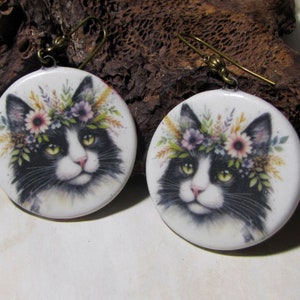 May include: A pair of round dangle earrings featuring a black and white cat illustration with a colourful floral crown on its head. The earrings have a white background and are attached to bronze-toned metal hooks for pierced ears.
