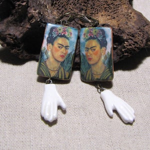 May include: A pair of rectangular earrings featuring a portrait of Frida Kahlo with a floral crown, suspended from bronze-toned hooks. Each earring includes a dangling white ceramic hand charm attached to the bottom of the rectangular portrait piece.
