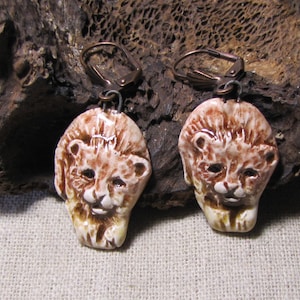 Puede incluir: Un par de pendientes de cerámica hechos a mano con caras de león. Los pendientes tienen un esquema de color marrón claro y crema con rasgos faciales detallados y alambres para las orejas de color cobre. Las caras de león están esculpidas con melenas texturizadas.