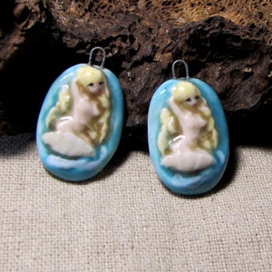 May include: Two oval-shaped ceramic pendants featuring a raised relief of a blonde figure with long hair, seated within a light blue water-like setting. Each pendant includes a small silver-toned metal loop at the top for attachment to a chain.
