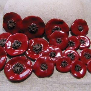Op de afbeelding: Een verzameling glanzende, rode keramische knopen in de vorm van klaprozen met donkerbruine centra. De knopen variëren in grootte en zijn gerangschikt op een neutraal gekleurd stoffen oppervlak.