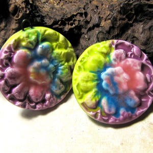 Peut inclure: Deux boucles d'oreilles rondes et colorées avec un motif floral. Les boucles d'oreilles présentent un effet tie-dye avec des nuances de violet, rose, bleu et jaune. Elles sont en céramique et ont une surface texturée. Elles sont présentées sur un fond sombre et texturé.