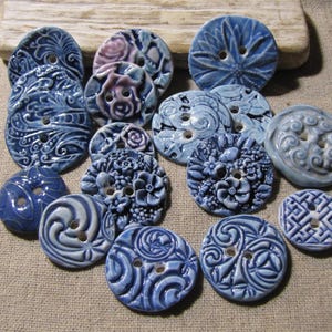 Op de afbeelding: Een verzameling ronde keramische knopen in verschillende tinten blauw en wit. Elke knoop heeft unieke reliëfpatronen, waaronder bloemmotieven, wervelende lijnen, geometrische vormen en ingewikkelde texturen. De knopen hebben twee gaatjes.