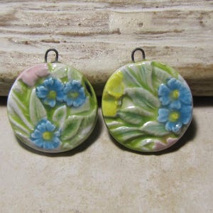 Pode incluir: Dois pingentes redondos de cerâmica com um design em relevo de flores azuis com centros amarelos, rodeadas por folhas verdes e pequenos detalhes em rosa. Cada pingente possui uma pequena argola de metal no topo para fixação.