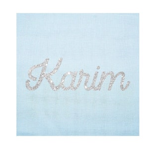 Peut inclure: Tissu bleu clair avec le nom "Karim" écrit en paillettes argentées.
