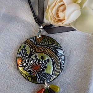 Puede incluir: Un collar negro con un colgante redondo que presenta un diseño floral colorido. El colgante tiene un acabado plateado y está acentuado con detalles verdes, dorados y rojos. El collar está adornado con un racimo de pequeñas cuentas de colores.