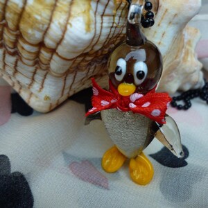 May include: A glass penguin charm with a red bow and white polka dots, hanging from a black beaded necklace. The charm is sitting on a white fabric with a seashell in the background.