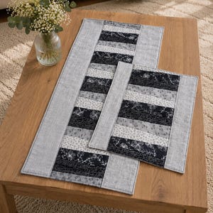 Könnte beinhalten: Ein rechteckiger Tischläufer und ein passendes kleineres rechteckiges Platzset, beide mit einem Patchwork-Design aus abwechselnden grauen Stoffrändern und einem Mittelteil aus horizontalen Streifen mit schwarz-weiß-grauen Blumenmustern.