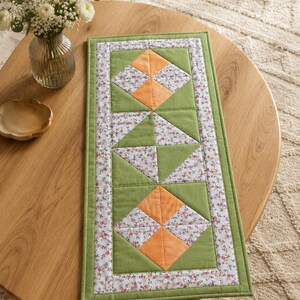 Könnte beinhalten: Ein rechteckiger, gesteppter Tischläufer mit einem Patchwork-Design aus salbeigrünem Stoff, orangefarbenen quadratischen Akzenten und einem weißen Rand mit kleinen rosa und violetten Blumenmotiven, abgeschlossen mit einem einfarbig grünen Außenrand.