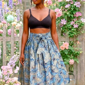 Op de afbeelding: Een vrouw draagt een zwarte bh en een knielange rok met een blauwe en gouden bloemenprint en een strik in de taille. De achtergrond toont een houten hek en diverse kleurrijke bloemen.