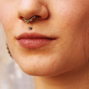 Puede incluir: Primer plano de la cara de una persona con un piercing en el septum y un piercing en el labio inferior. El piercing en el septum tiene un anillo de cuentas doradas, y el piercing en el labio inferior tiene una pequeña piedra preciosa azul.