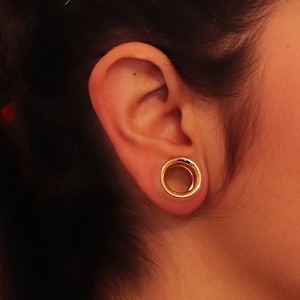 May include: A close-up of a person's ear with a gold-colored ear gauge. The gauge is a large ring with a smooth, polished finish.