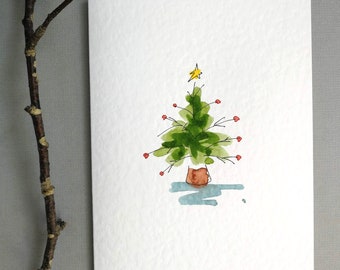May include: A watercolor Christmas card featuring a whimsical green Christmas tree with red ornaments and a yellow star on top. The tree is in a brown pot, with a blue shadow beneath it. The card is on a white background.