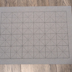 May include: A gray calligraphy practice sheet with a grid pattern. The sheet has a rectangular shape and features a series of squares and diagonal lines, designed to guide the user in writing characters. The sheet is placed on a wooden surface.