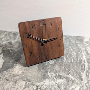 Puede incluir: Un pequeño reloj cuadrado de madera con manecillas negras y números romanos. El reloj está hecho de madera marrón oscura y tiene un diseño simple y minimalista.