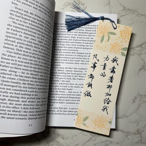 Puede incluir: Un marcador de página de papel con un patrón floral y caligrafía japonesa. El marcador tiene un borla azul. El texto en el marcador dice "我靠著加给我力童心凡事布食做"