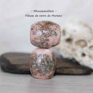 Puede incluir: Dos cuentas de vidrio hechas a mano apiladas una encima de la otra. Las cuentas son de color rosa claro con manchas marrones y verde azulado. La cuenta superior tiene forma de cubo y la inferior es ovalada. El texto "- SRousseauGlass - Fileuse de verre de Murano" está en la parte superior.