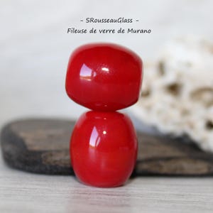 Puede incluir: Cuenta de cristal de Murano roja de dos piezas. La pieza superior es cuadrada y la inferior redondeada. El cristal es brillante y refleja la luz. El texto "- SRousseauGlass - Fileuse de verre de Murano" está en la imagen.