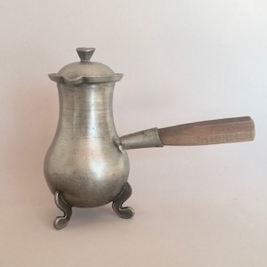 Può includere: Una caffettiera in metallo argentato con un manico in legno e tre gambe. La caffettiera ha un coperchio con un piccolo pomo in cima.