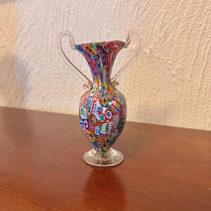 May include: A small, colorful glass vase with two handles. The vase is decorated with a mosaic of small, colorful glass pieces. The vase is on a wooden surface.