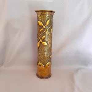 May include: A tall, cylindrical brass vase with a textured surface and gold leaf accents. The vase features a detailed floral design and is approximately 25 cm tall. The top and bottom rims have a decorative edge.