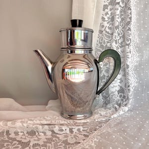 Puede incluir: Una cafetera cromada vintage con asa y tapa negras. La cafetera tiene una superficie reflectante y el asa es de color verde oscuro. La cafetera está sobre un mantel de encaje blanco.
