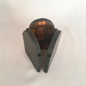May include: A small, gray, stone-like box with a hinged lid. The lid is open, revealing a gold-colored image of Jesus inside.
