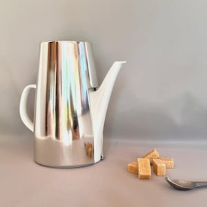 Könnte beinhalten: Eine glänzende, silberfarbene Kaffeekanne mit weißem Ausguss und Griff. Daneben liegen mehrere braune Würfelzucker und ein kleiner Löffel. Die Kanne spiegelt die Umgebung wider.
