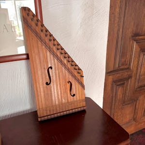 May include: A wooden instrument with a triangular form, with strings and tuning pegs along the top. It has two f-shaped sound holes. The instrument is on a dark brown surface. The wood has a warm, natural tone, similar to a traditional autoharp.