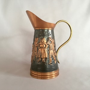 Op de afbeelding: Vintage koperen en messing kan met een decoratief reliëf van figuren in traditionele kleding. De kan heeft een koperen tuit en voet, een donkergrijze body en een messing handvat. Een uniek stuk.