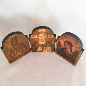 May include: A three-panel wooden icon set with gold-toned images of Jesus, a king, and an angel. The panels are hinged and can be folded together.
