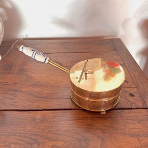 Peut inclure: Une boîte ronde en laiton et en céramique avec un couvercle à charnière et une poignée en céramique blanche et bleue. La boîte a un motif décoratif sur le couvercle et repose sur une surface en bois.