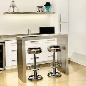 Op de afbeelding: Een wit keuken eiland met een stalen aanrechtblad en twee industriële barstoelen. Het eiland heeft een ingebouwde wijnkoeler en een gootsteen. Boven het aanrechtblad is een plank met decoratieve items.