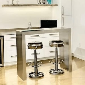 Puede incluir: Una barra de cocina moderna con una encimera de acero inoxidable y gabinetes blancos. Dos taburetes de bar de estilo industrial con asientos de metal y una base en forma de tambor están colocados frente a la barra. La barra tiene una nevera de vino incorporada.