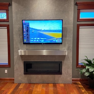 Puede incluir: Una chimenea moderna con un hogar negro y una repisa de acero inoxidable. Un televisor de pantalla plana está montado sobre la chimenea. El televisor muestra un coche de carreras en una pista con la palabra "ally" en la esquina inferior derecha.