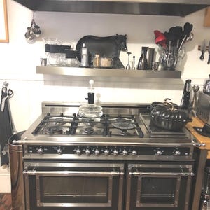 May include: A black and silver professional range cooker with six burners, two ovens, and a warming drawer. The cooker has a stainless steel hood above it.