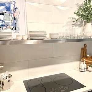 Puede incluir: Escena de cocina moderna con una placa de cocción de vidrio negro con cuatro quemadores. Platos, cuencos y vasos blancos están dispuestos en un estante de acero inoxidable. Una pintura abstracta enmarcada y un jarrón con vegetación decoran.