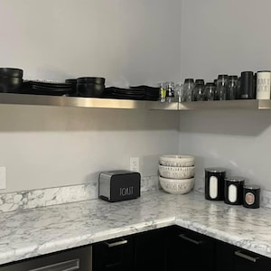 May include: A white countertop with a black toaster labelled "TOAST" and three white ceramic bowls with black lettering. The bowls are stacked on top of each other. There are also four glass jars with black lids on the counter. The countertop is next to a stainless steel shelf with black bowls and plates on it.