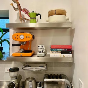 Puede incluir: Dos estantes de acero inoxidable con una variedad de artículos de cocina, que incluyen una máquina de espresso naranja brillante, una cafetera verde y blanca, un vaporizador de cerámica blanco, una pila de libros de cocina y un juego de taza y platillo con estampado en blanco y negro.