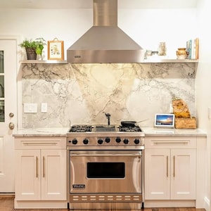 Puede incluir: Una campana extractora de acero inoxidable con un salpicadero de mármol blanco y armarios blancos. La campana extractora está sobre una cocina de acero inoxidable con un horno y cuatro quemadores. La cocina está sobre una encimera blanca con un fregadero y un grifo blancos.