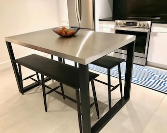 Könnte beinhalten: Ein Küchentisch aus Edelstahl mit einem schwarzen Metallrahmen und zwei schwarzen Holzbänken. Auf dem Tisch steht eine Schüssel mit Obst. Der Tisch steht in einer Küche mit weißen Fliesenboden und einem blau-weiß gemusterten Teppich.