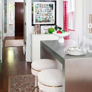 May include: A white kitchen island with a stainless steel worktop and two white upholstered bar stools with gold trim. The island is in a kitchen with white cabinets, a red and white patterned curtain, and a framed print of a plant collection.