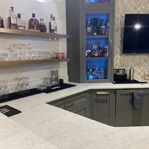 May include: A home bar with a gray cabinet with glass doors and shelves filled with bottles of liquor. The cabinet is next to a white countertop with a black mat and a row of glasses. The countertop is also next to a gray cabinet with a black towel hanging on the door. The countertop is made of white quartz.