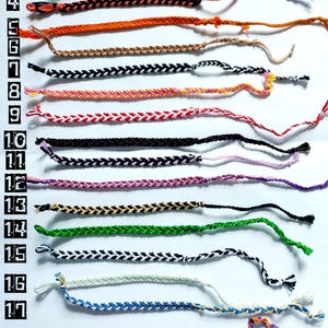 May include: A collection of nineteen colorful friendship bracelets, each with a unique woven pattern. The bracelets vary in color, including blue, green, orange, black, and white. Each bracelet is approximately 6 inches long.