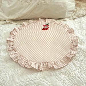 Può includere: Un cuscino rotondo con motivo a quadretti vichy rosa chiaro e bianco, caratterizzato da un bordo arricciato e da un piccolo motivo a ciliegia rossa ricamato con gambo e foglia verdi al centro della superficie superiore.