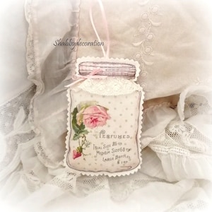 Può includere: Un ornamento in tessuto a forma di barattolo con illustrazione di rosa, bordo in pizzo bianco e nastro rosa. Il testo recita: Perfumes, Trial Size 25 cts, Medium Size 50 cts, Large Bottles $1.00. Lo sfondo presenta un motivo a pois chiaro.