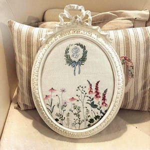 Può includere: Ricamo ovale con cornice bianca con un motivo floreale. Il ricamo include una ghirlanda con una lettera, un fiocco blu e vari fiori ricamati in rosa, viola e rosso. La cornice ha un fiocco decorativo nella parte superiore.