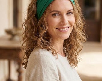 May include: A woman with curly brown hair wearing a green knotted headband and a light beige long-sleeved shirt. The headband is a solid, vibrant green color. The woman is smiling, and the background is blurred.